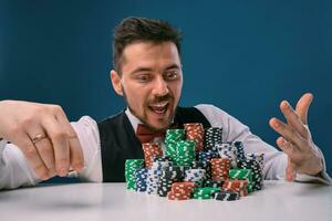 Man in black vest and shirt sitting at white table with stacks of chips on it, posing on blue studio background. Gambling, poker, casino. Close-up. photo