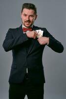 hombre en negro clásico traje y rojo corbata de moño demostración dos jugando tarjetas mientras posando en contra gris estudio antecedentes. juego, póker, casino. de cerca. foto