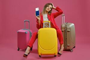 rubia mujer en rojo traje pantalón, blanco blusa, negro tacones ella sonriente, sentado entre vistoso maletas, participación pasaporte y boleto, rosado antecedentes foto