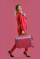 rubia hembra en rojo traje pantalón, alto negro tacones ella sonriente, que lleva maleta, participación pasaporte y boleto, rosado antecedentes. de viaje concepto foto