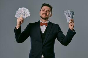 Man in black suit and red bow-tie is showing two fans of hundred dollar bills, posing on gray studio background. Gambling, poker, casino. Close-up. photo