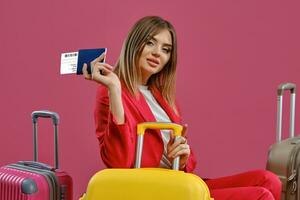 Blonde girl in red pantsuit, white blouse. She is smiling while sitting among colorful suitcases, holding passport and ticket, pink background photo