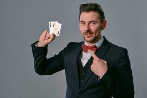 Man in black classic suit and red bow-tie showing two playing cards while posing against gray studio background. Gambling, poker, casino. Close-up. photo