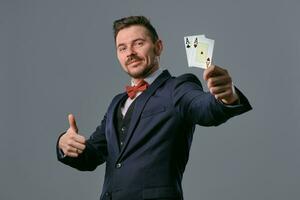 Man in black classic suit and red bow-tie showing two playing cards while posing against gray studio background. Gambling, poker, casino. Close-up. photo