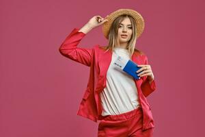 Blonde female in straw hat, white blouse and red pantsuit. She is holding passport and ticket while posing against pink studio background. Close-up photo