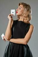 Blonde girl in black stylish dress showing two playing cards, posing against gray background. Gambling entertainment, poker, casino. Close-up. photo