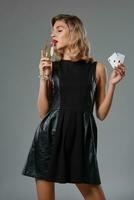 Blonde girl in black dress showing two playing cards, drinking champagne, posing against gray studio background. Gambling, poker, casino. Close-up. photo