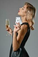 Blonde girl in black dress holding two playing cards and glass of champagne, posing against gray background. Gambling, poker, casino. Close-up. photo