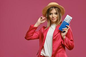 Blonde girl in straw hat, white blouse and red pantsuit. She is holding passport and ticket while posing against pink studio background. Close-up photo