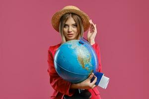 Woman in straw hat, white blouse and red pantsuit. She is touching headdress, holding globe, passport and ticket, posing on pink background. Close-up photo