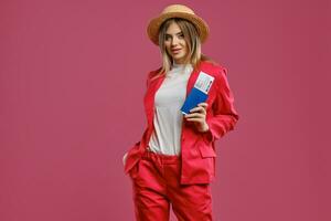 Blonde lady in straw hat, white blouse and red pantsuit. She is holding passport and ticket while posing against pink studio background. Close-up photo