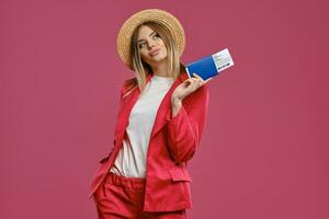 Blonde woman in straw hat, white blouse and red pantsuit. She is holding passport and ticket while posing against pink studio background. Close-up photo