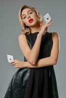 Blonde girl in black leather dress showing two playing cards, posing against gray background. Gambling entertainment, poker, casino. Close-up. photo