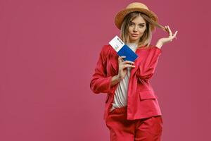 Blonde woman in straw hat, white blouse and red pantsuit. She is holding passport and ticket while posing against pink studio background. Close-up photo