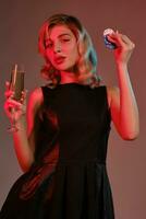 Blonde girl in black dress holding glass of champagne and chips, posing against colorful background. Gambling, poker, casino. Close-up. photo