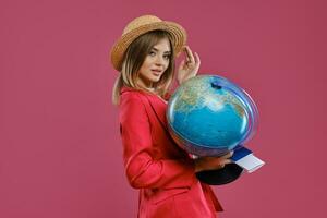 Blonde lady in straw hat, white blouse and red pantsuit. She is holding a globe, passport and ticket, posing sideways on pink background. Close-up photo