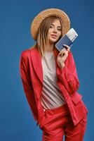 Blonde girl in straw hat, white blouse and red pantsuit. She smiling, showing passport and ticket while posing on blue studio background. Close-up photo