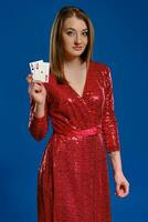 Blonde woman with make-up, tattooed hand, in red sequin dress is showing two aces, posing on blue background. Gambling, poker, casino. Close-up. photo
