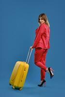 Blonde woman in red pantsuit, high black heels. She has raised her leg, posing back to the camera with yellow suitcase on blue background. Full length photo