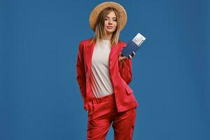 Blonde lady in straw hat, white blouse and red pantsuit. She smiling, holding passport and ticket while posing on blue studio background. Close-up photo