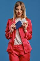 Blonde woman in white blouse and red pantsuit. She smiling, holding passport and ticket while posing on blue studio background. Close-up photo