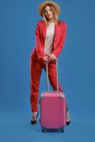 Blonde woman in straw hat, white blouse, red pantsuit, high black heels. She is leaning on a handle of pink suitcase while posing on blue background photo