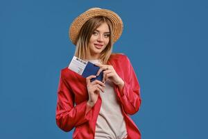 rubia dama en Paja sombrero, blanco blusa, rojo traje pantalón. ella es participación pasaporte y boleto, posando en azul antecedentes. de viaje concepto. de cerca foto