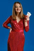 Blonde lady with make-up, in red sequin dress is smiling, showing two aces, posing on blue background. Gambling, poker, casino. Close-up. photo