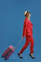 Blonde female in hat, red pantsuit, high heels. She carrying pink suitcase by handle, walking on blue background. Side view, full length photo