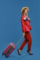 Blonde model in hat, white blouse, red pantsuit, high heels. Carrying pink suitcase, showing passport and ticket, posing sideways on blue background photo