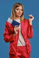 Blonde female in white blouse and red pantsuit. She smiling, holding passport and ticket while posing on blue studio background. Close-up photo