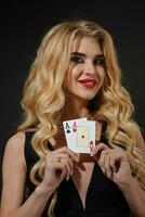 Blonde model in black stylish dress and necklace. She smiling, showing two aces, posing on black background. Poker, casino. Close-up photo