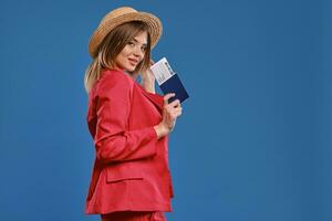 Blonde woman in straw hat, white blouse, red pantsuit. She is holding passport and ticket, posing sideways on blue background. Travelling. Close-up photo