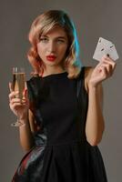 Blonde girl in black dress holding two playing cards and glass of champagne, posing against gray background. Gambling, poker, casino. Close-up. photo