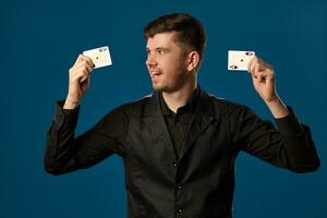 Newbie in poker, in black vest and shirt. Holding two playing cards while posing against blue studio background. Gambling, casino. Close-up. photo