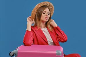 Blonde model in straw hat, white blouse, red pantsuit. Sitting leaning on pink suitcase, posing on blue background. Travelling concept. Close-up photo