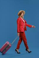 Blonde woman in hat, white blouse, red pantsuit, high heels. She carrying pink suitcase by handle, walking on blue background. Side view, full length photo