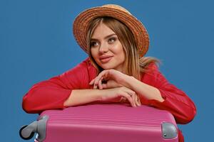 Blonde model in straw hat and red jacket. She is smiling while sitting leaning on pink suitcase, posing on blue studio background. Close-up photo