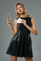 Blonde girl in black dress holding two playing cards and glass of champagne, posing against gray background. Gambling, poker, casino. Close-up. photo