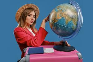 Blonde female in straw hat, white blouse, red jacket. Holding globe standing on pink suitcase, passport and ticket nearby, posing on blue background photo