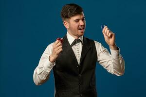 Newbie in poker, in black vest and white shirt. Holding some colored chips. Posing against blue background. Gambling, casino. Close-up. photo