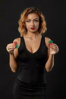 Ginger girl wearing dark dress is posing holding red and green chips in her hands standing against black studio background. Casino, poker. Close-up. photo