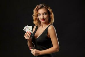 Ginger girl in a dark dress posing holding playing cards and chips in her hands standing against a black studio background. Casino, poker. Close-up. photo