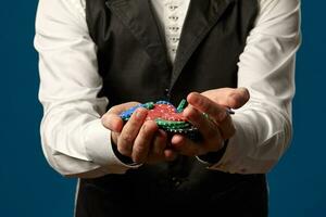 Noob in poker, in black vest and white shirt. Holding some colored chips. Posing against blue background. Gambling, casino. Close-up. photo