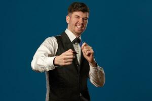 Newbie in poker, in black vest and white shirt. Holding some colored chips. Posing against blue background. Gambling, casino. Close-up. photo