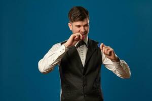 Noob in poker, in black vest and white shirt. Holding some colored chips. Posing against blue background. Gambling, casino. Close-up. photo