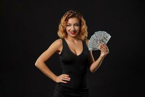 Ginger girl in dark dress posing holding fan of hundred-dollar bills in her hands standing against a black studio background. Casino, poker. Close-up. photo