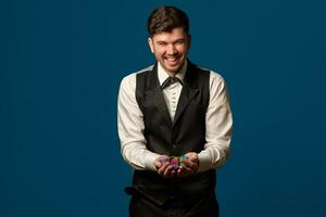 Newbie in poker, in black vest and trousers, white shirt. Holding some colored chips. Posing against blue background. Gambling, casino. Close-up. photo