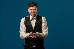 Noob in poker, in black vest and trousers, white shirt. Holding some colored chips. Posing against blue background. Gambling, casino. Close-up. photo