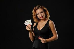 Ginger girl in a dark dress posing holding playing cards and chips in her hands standing against a black studio background. Casino, poker. Close-up. photo